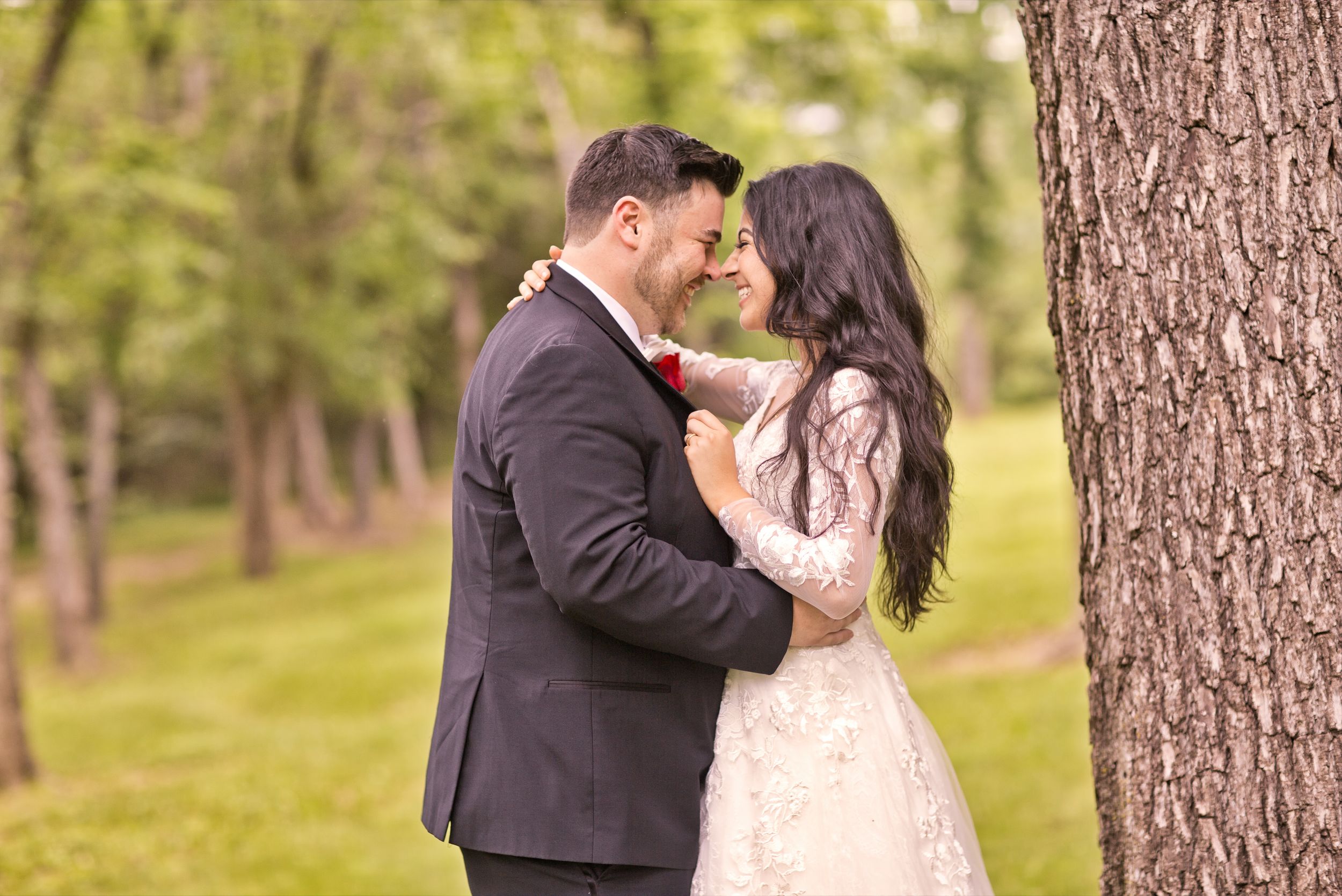Bride and groom nearly kissing beside a large tree in a serene green park setting.