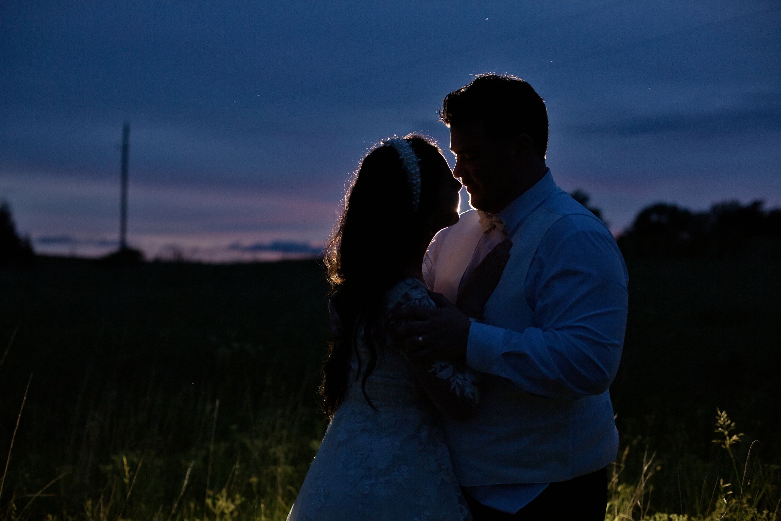 Romantic silhouette of bride and groom embracing in a field beneath a stunning purple twilight sky.