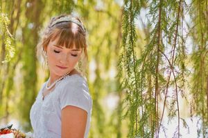 Elegant bride in white dress standing under weeping willow branches in soft sunlight.