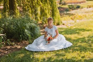 Bride in white gown sitting on grass holding colorful bouquet under a weeping willow tree.