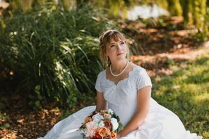 Bride in white gown holding peach bouquet sits outdoors among lush green foliage on her wedding day.