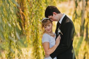 Groom tenderly kisses bride's forehead under weeping willow branches in romantic outdoor wedding portrait.