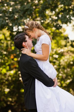 Groom lifts bride in white wedding gown for a romantic kiss surrounded by lush green trees.