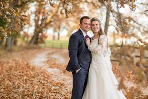 Smiling bride and groom pose together on a leaf-covered country path in autumn wedding portrait.