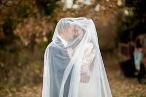 Bride and groom share a romantic moment hidden beneath bride's flowing veil in autumn woodland setting.