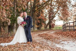 Bride holding red bouquet and groom pose on a rustic leaf-lined path in fall wedding portrait.