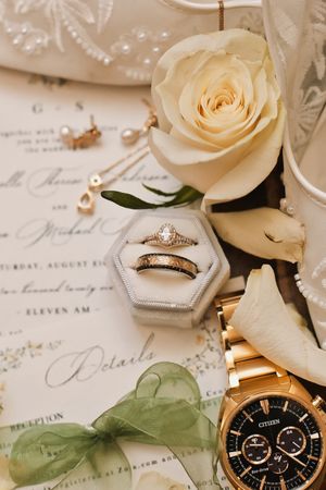 Wedding flat lay featuring diamond rings in velvet box, white rose, Citizen watch, and invitation.