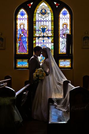 Bride and groom sharing intimate moment before colorful stained glass windows inside historic church.