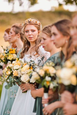 Close-up of bride wearing floral crown standing with bridesmaids holding white and yellow bouquets.