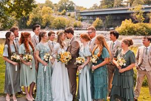 Wedding party in sage green and tan attire kissing and celebrating outdoors by riverside.