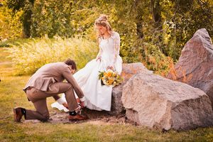 Groom kneeling to fix bride's shoe near large rocks in a sunlit outdoor wedding setting.