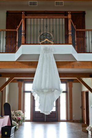 Elegant white wedding gown hanging on a wooden hanger in a rustic barn venue interior.