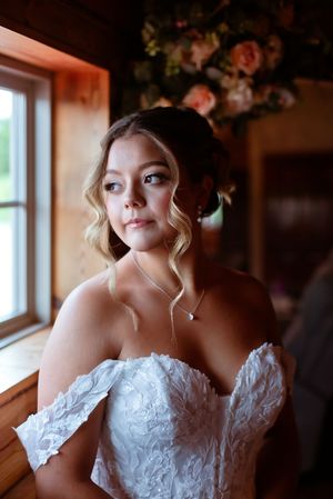 Beautiful blonde bride in off-shoulder lace gown gazes thoughtfully out a window with soft natural light.