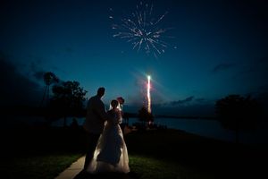 Bride and groom silhouetted against colorful fireworks display over lake at night.