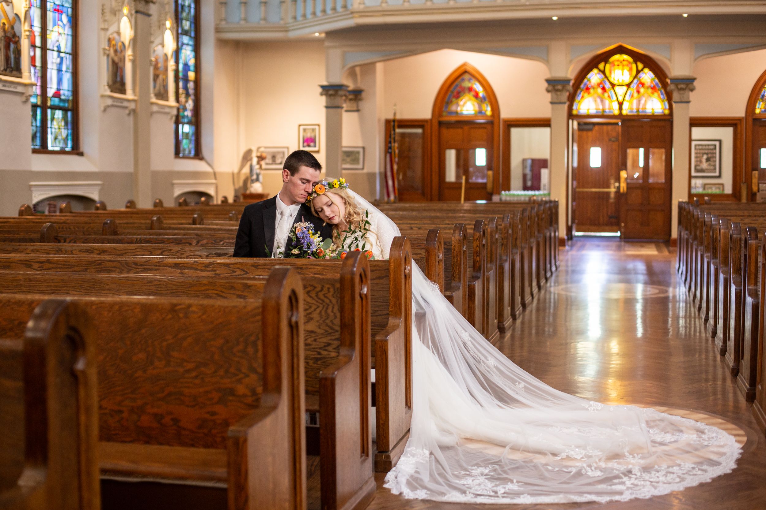 Bride and groom share a romantic kiss in a beautiful church aisle with wooden pews and stained glass windows.