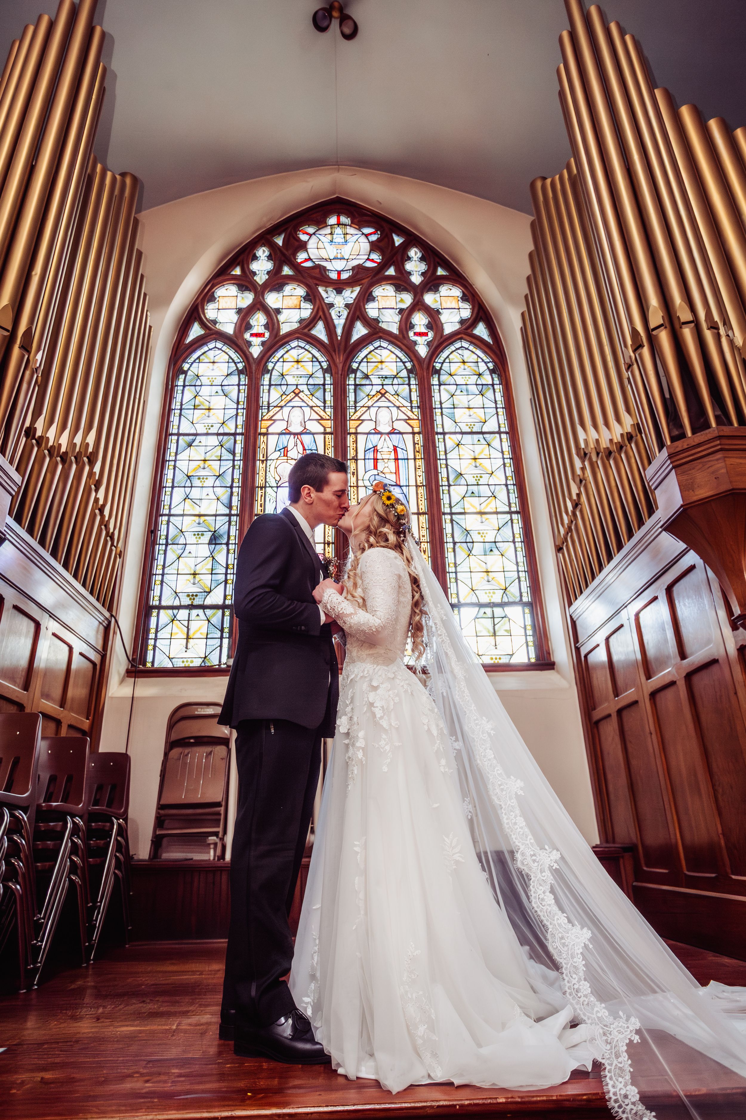 Bride and groom kiss at the altar beneath a stunning stained glass window and pipe organ in a historic church.
