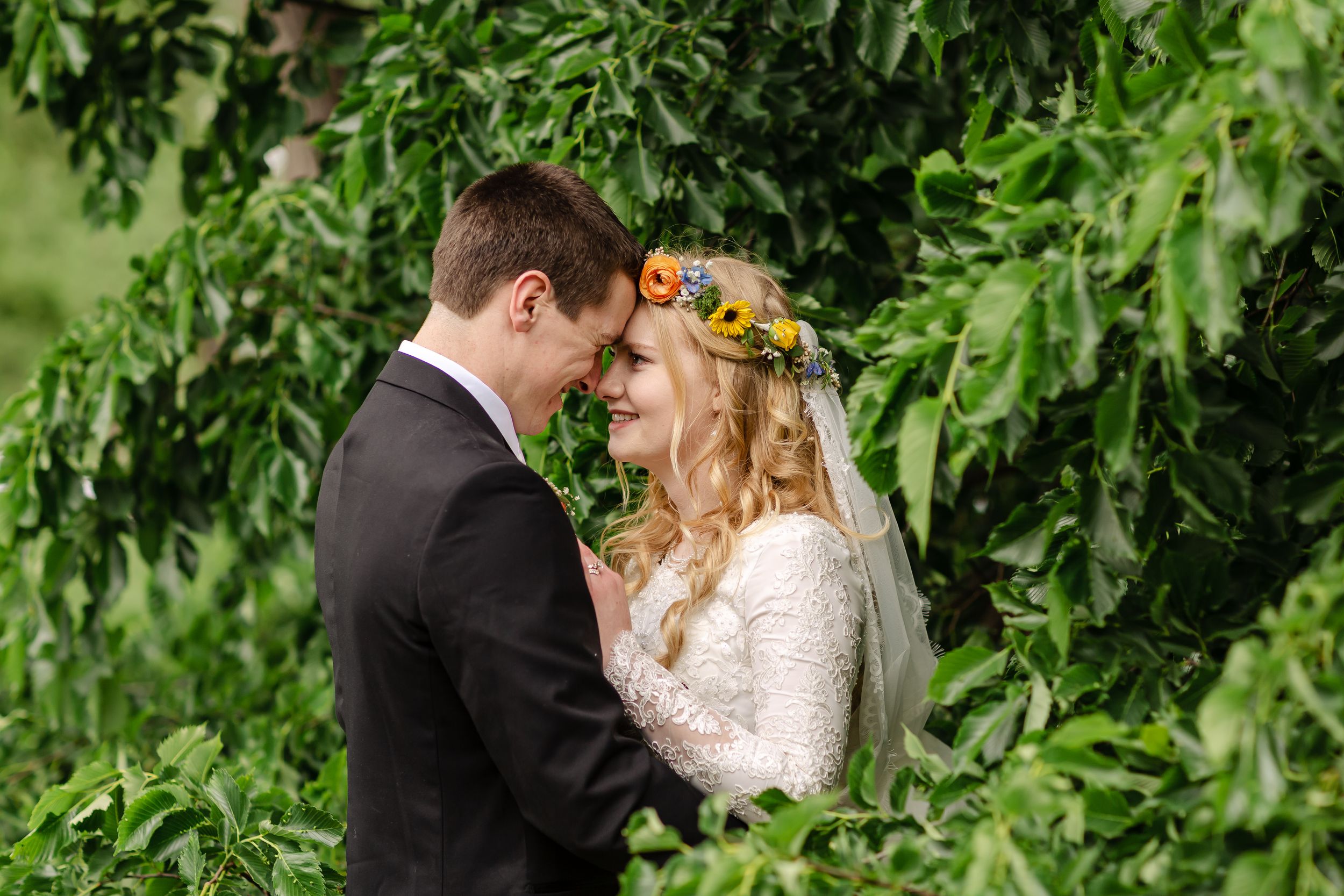 Bride wearing floral crown and groom share an intimate moment surrounded by lush green foliage outdoors.