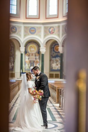 Bride and groom sharing romantic kiss in ornate church aisle with colorful frescoes and stained glass windows.
