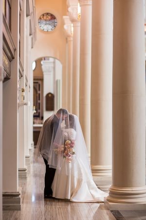 Bride and groom sharing a kiss under a flowing veil in an elegant church with tall white columns.