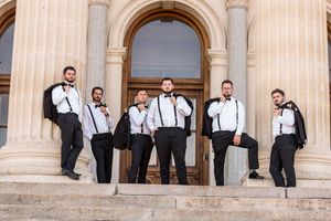 Six groomsmen in black tuxedos with suspenders pose on stone steps outside a grand building.