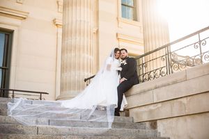 Bride in flowing veil and groom sitting together on stone steps of classical pillared building.