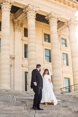 Bride and groom stand on grand stone steps of a neoclassical building with tall Corinthian columns on their wedding day.
