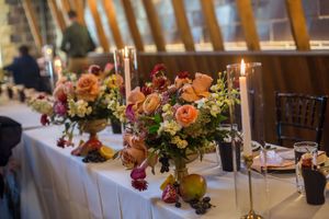 Elegant wedding reception table with lush floral centerpieces, candles, and fruit accents in a rustic barn venue.