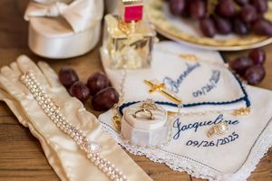 Wedding flat lay featuring gold rings on embroidered handkerchief, satin gloves, perfume, and grapes on wooden surface.