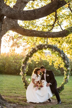 Bride and groom share a romantic moment inside a circular floral arch beneath a sunlit tree at golden hour.