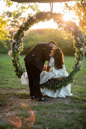 Bride and groom sharing a romantic kiss under a floral wreath swing at golden hour in a lush garden.