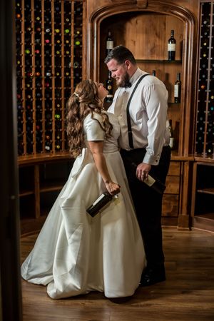 Bride and groom share a romantic moment in a wine cellar, holding bottles among wooden wine racks.