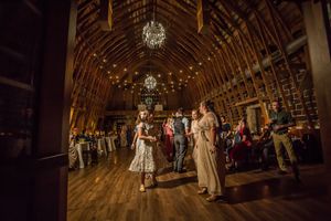 Guests dance and mingle inside a rustic barn wedding reception with warm chandelier lighting.
