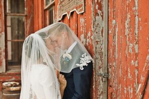 Bride and groom kissing under wedding veil against a weathered red barn door.