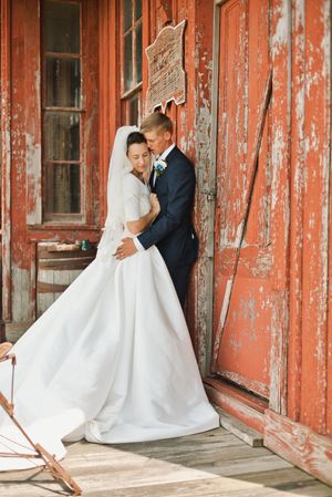 Bride and groom embracing beside a rustic red peeling barn wall on wooden porch.