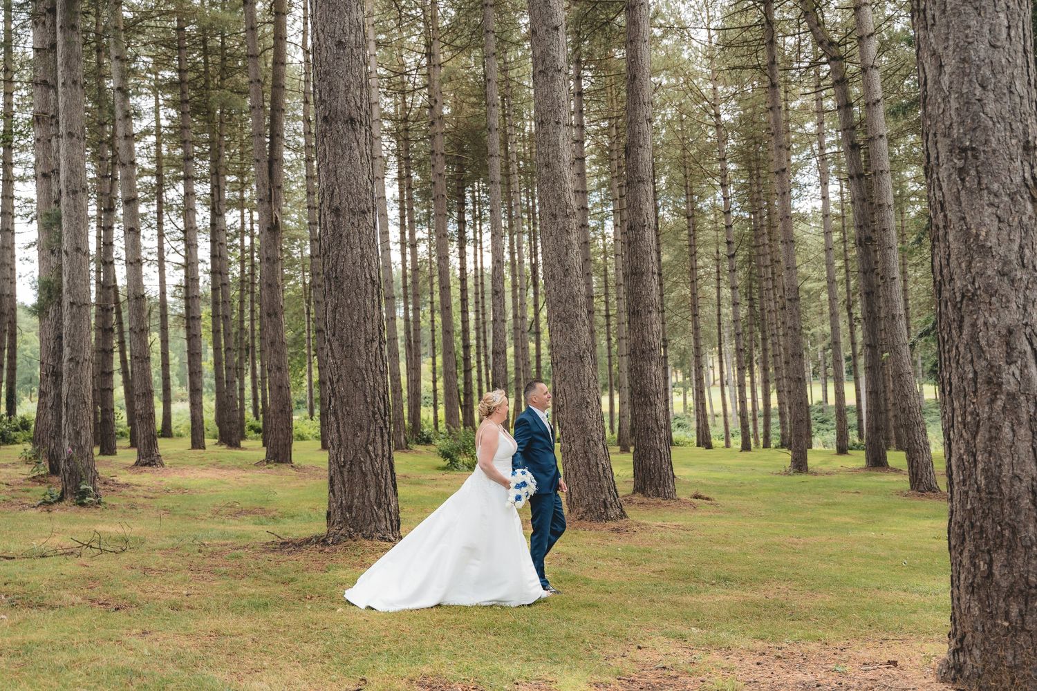 A wedding couple walks through a tall pine forest setting with sunlight filtering through the trees.