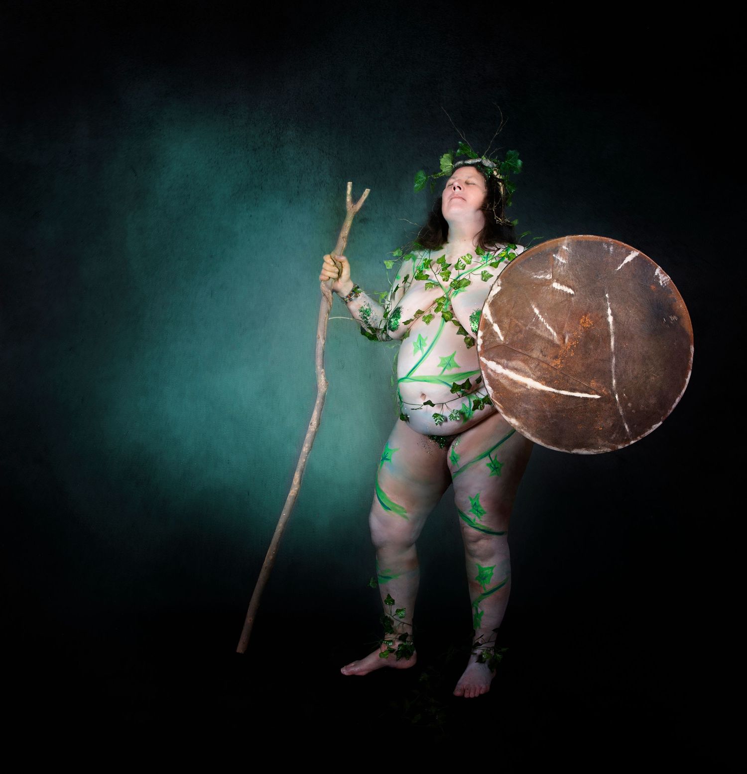 Celtic warrior figure in leaf-patterned costume holds wooden staff and shield against mystical teal backdrop.