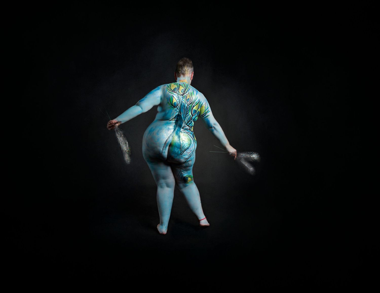 A performer in a blue and turquoise painted Peacock strikes a dramatic pose against a dark background.