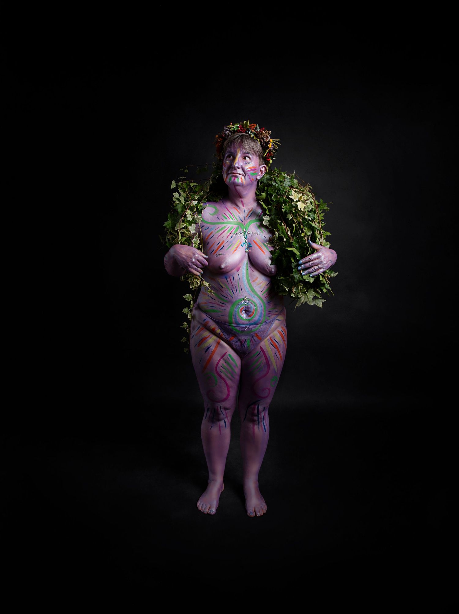 Figure with colorful body paint poses against dark background wearing floral crown and greenery around shoulders.