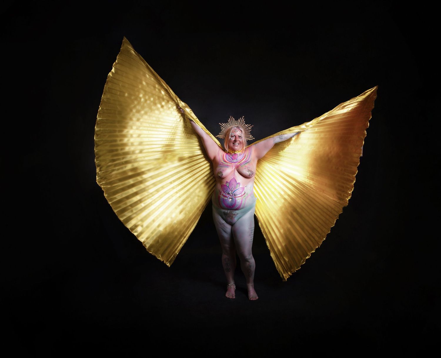 The Goddess in a pink top dances with large golden wing-like props against a dark background.