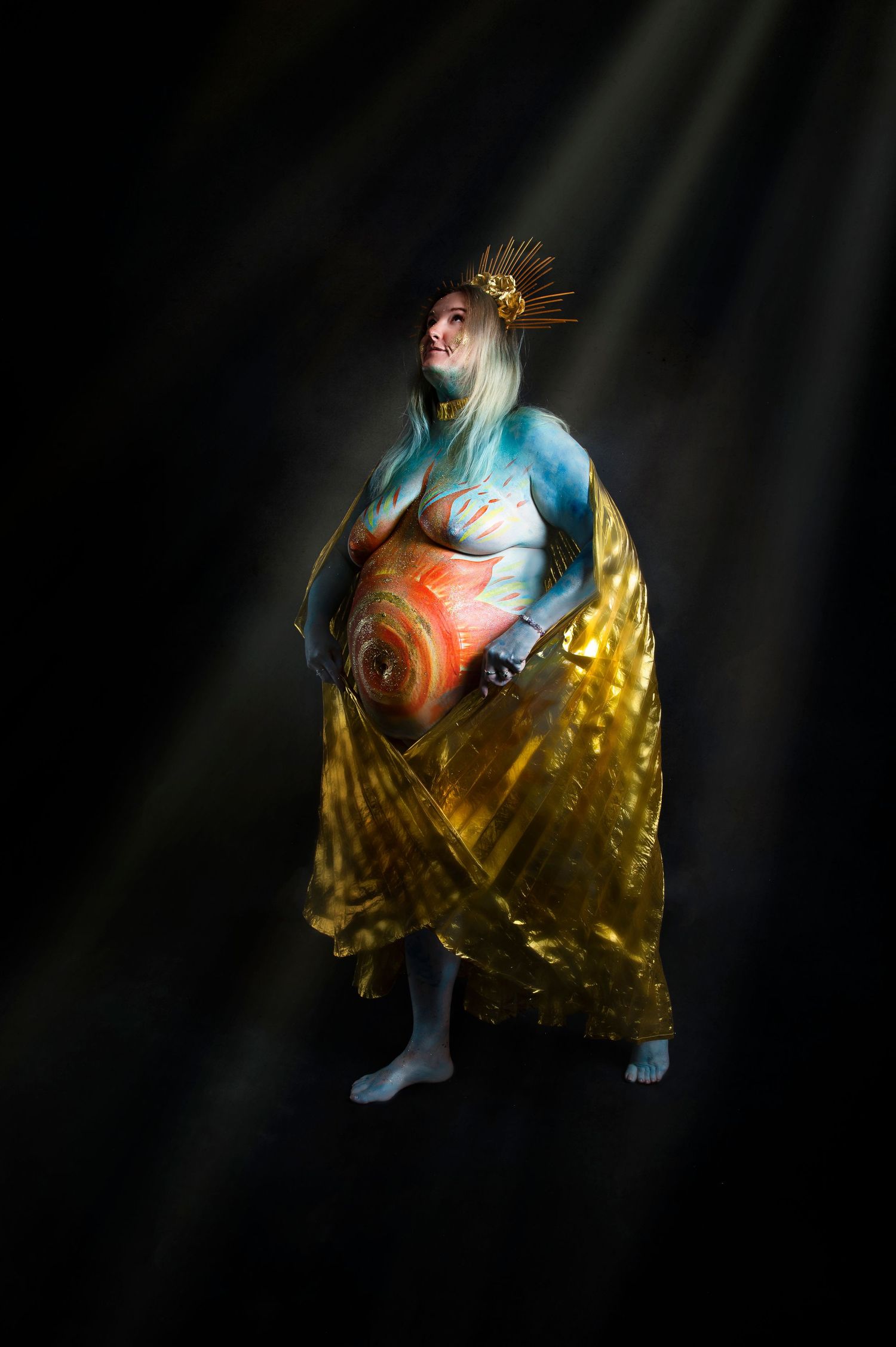 Maternity Goddess in a golden robe with blue-painted skin and crown stands dramatically illuminated by beams of light against darkness.