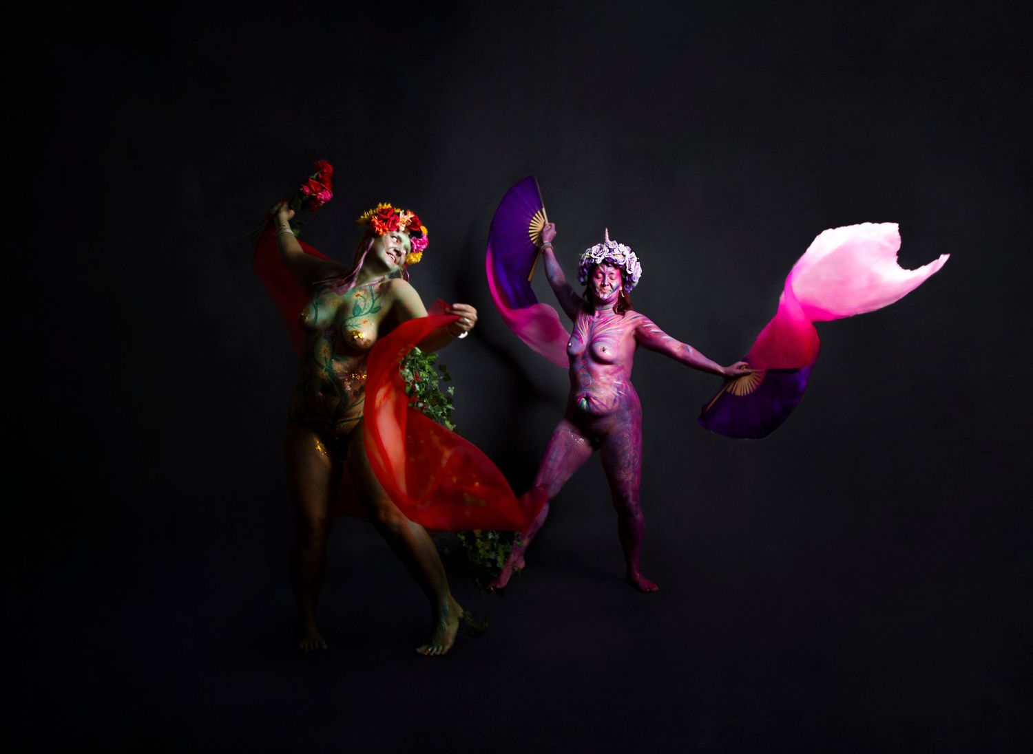 Two Goddess's in colorful theatrical costumes dance with flowing fabrics against a dark background.