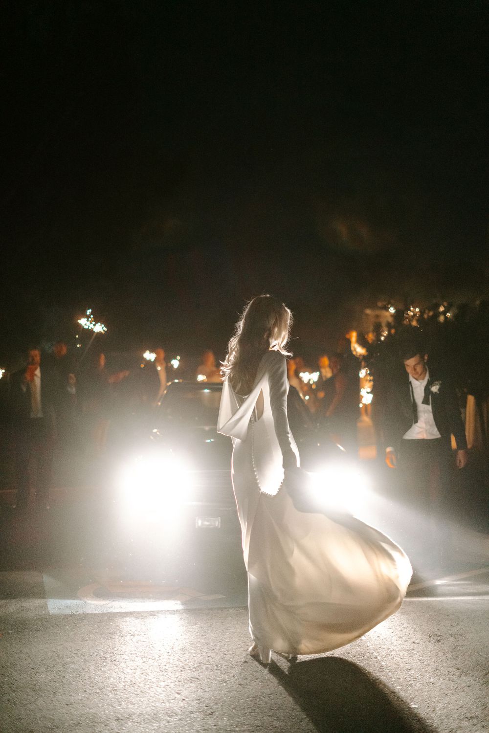 Silhouette of a bride dancing in a flowing dress illuminated by car headlights at night.
