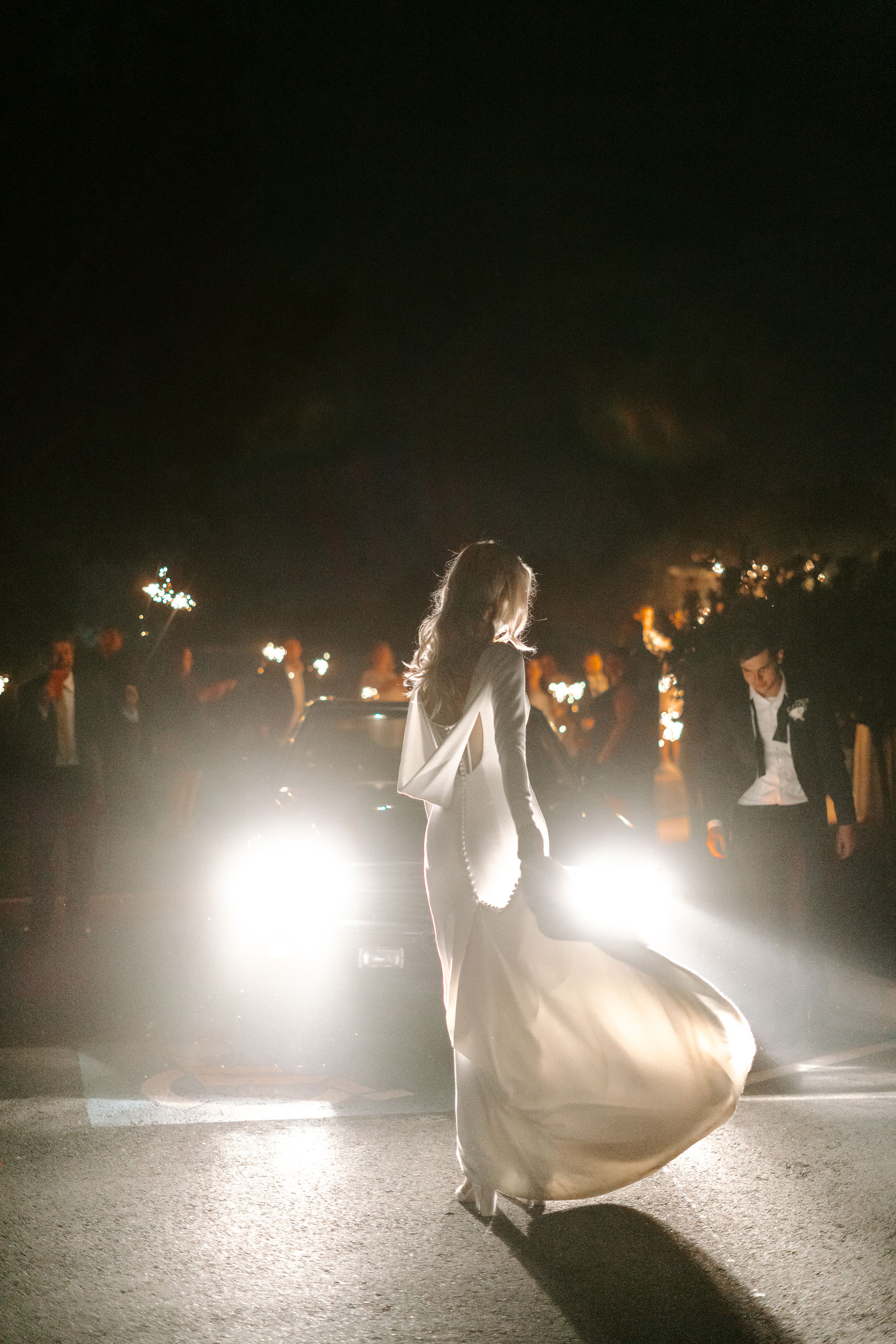 Silhouette of a bride dancing in a flowing dress illuminated by car headlights at night.