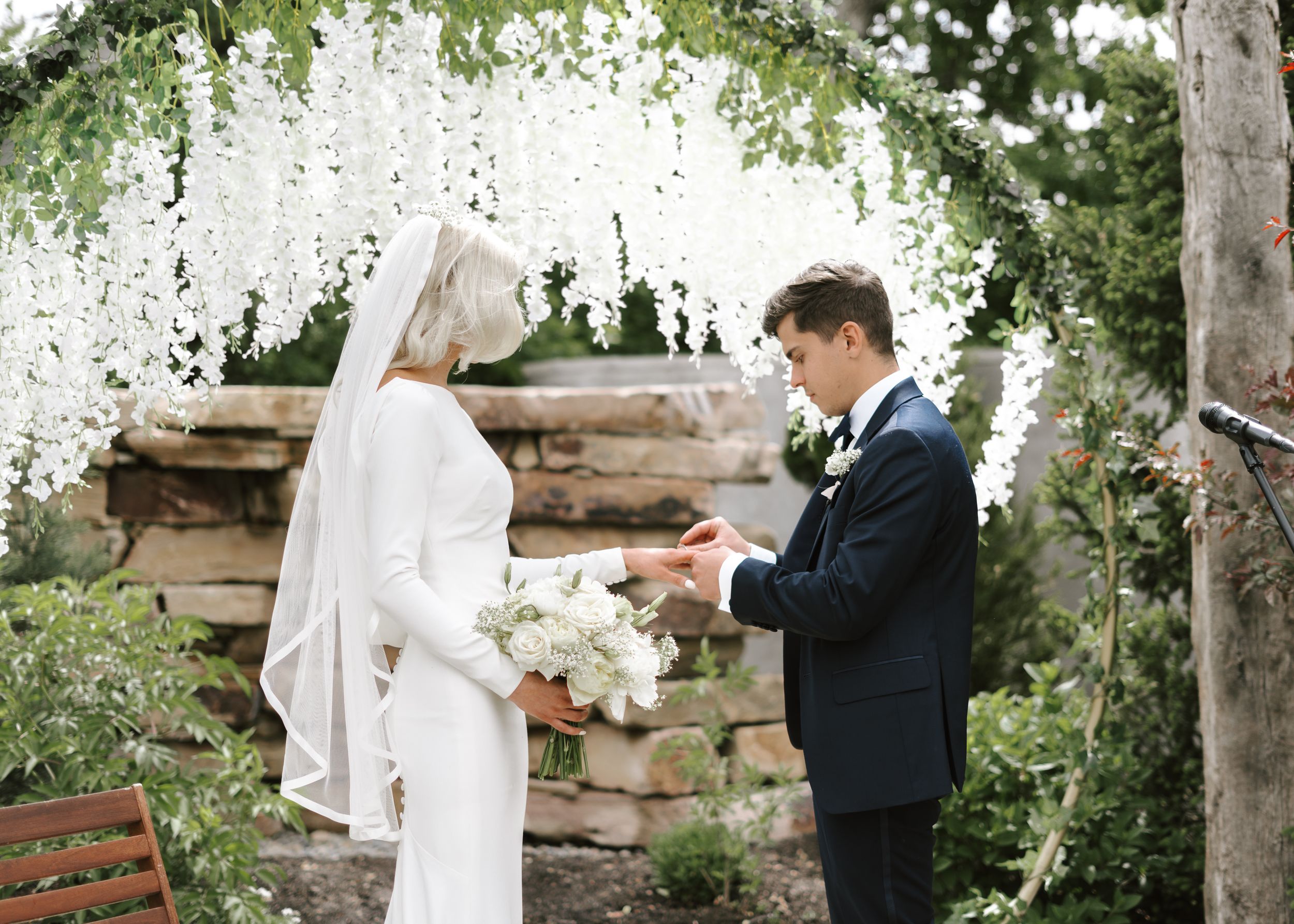 Wedding ceremony setup with white floral arch and wooden backdrop in an outdoor garden setting.