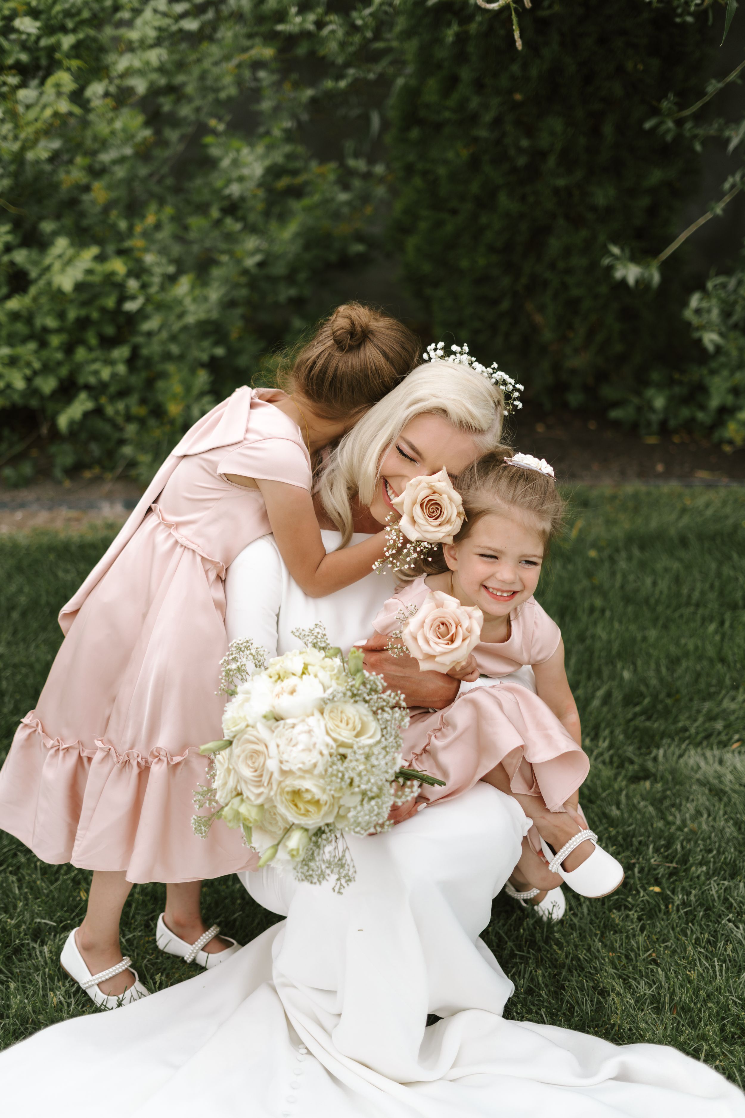 Young flower girls in pink dresses share a sweet moment with bouquets in a garden setting.
