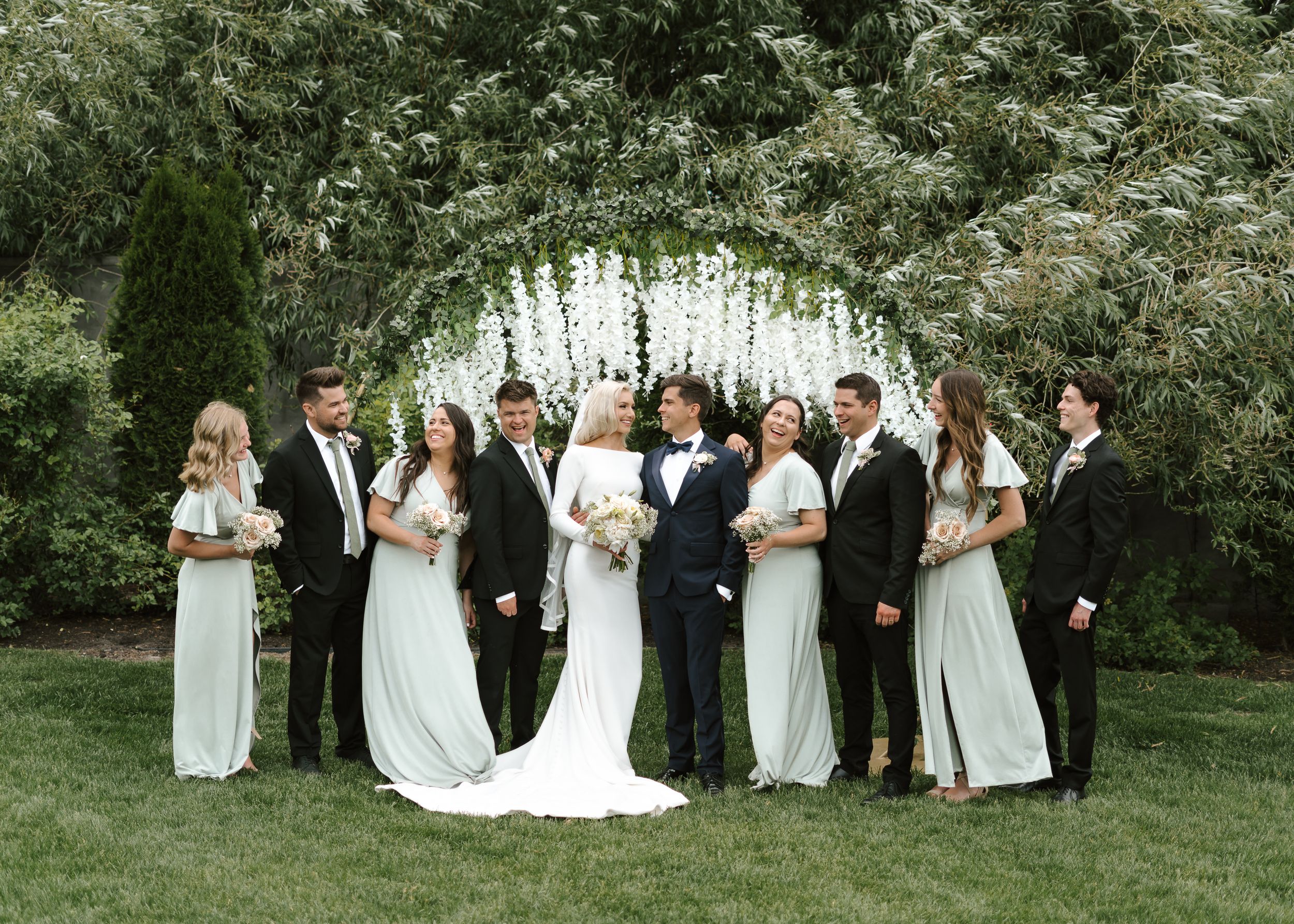 Wedding party poses together under a beautiful white floral arch in an outdoor garden setting.