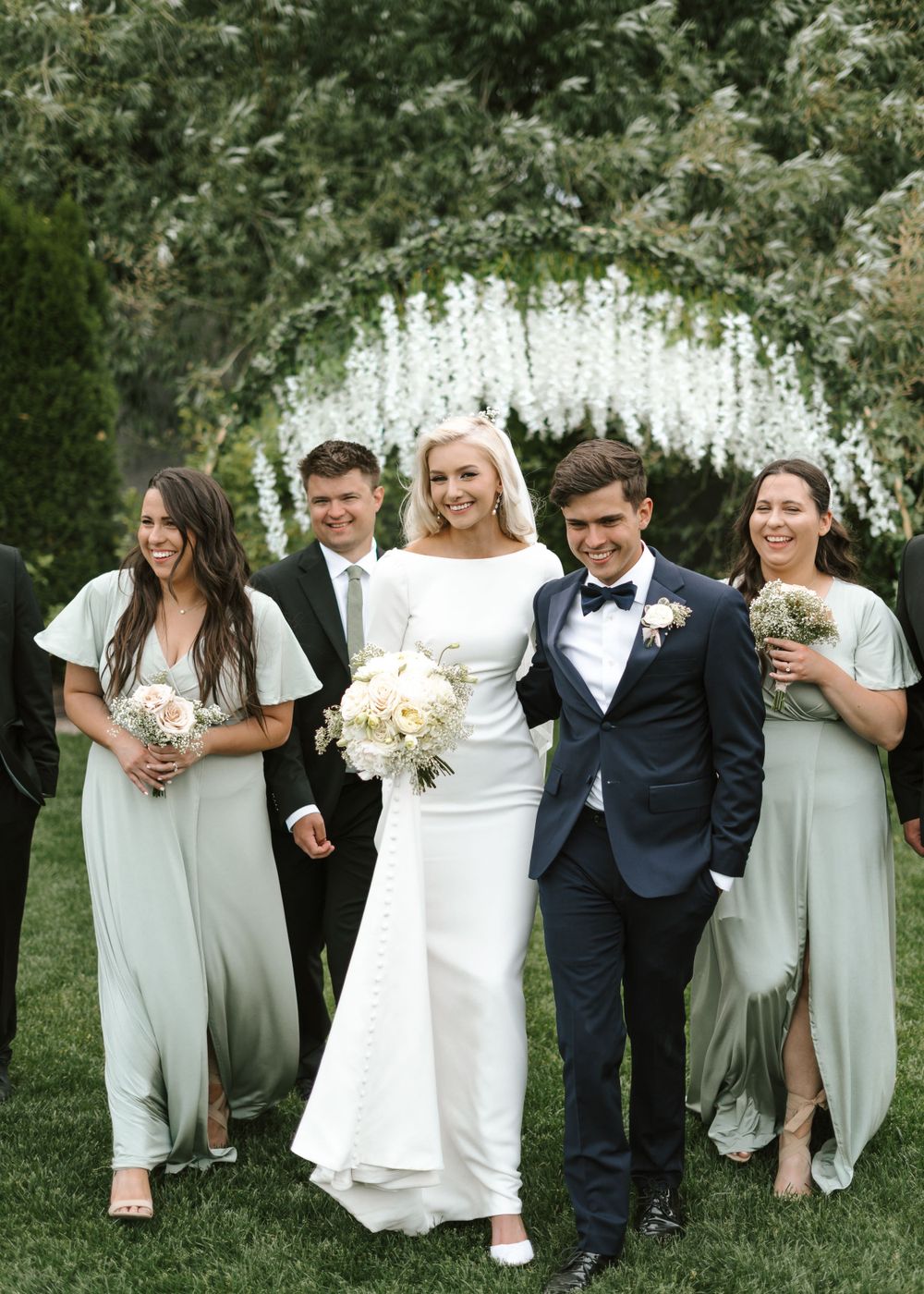 A wedding party walks and laughs together under a floral arch in a garden setting.