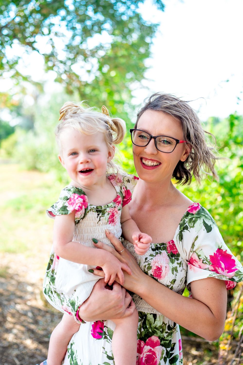 Mother and young child in matching floral dresses smile together in natural setting.