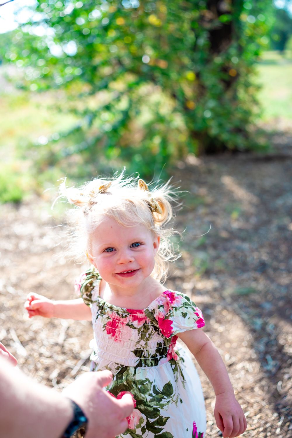 Smiling toddler in floral dress reaches out while playing outdoors in sunlit setting.