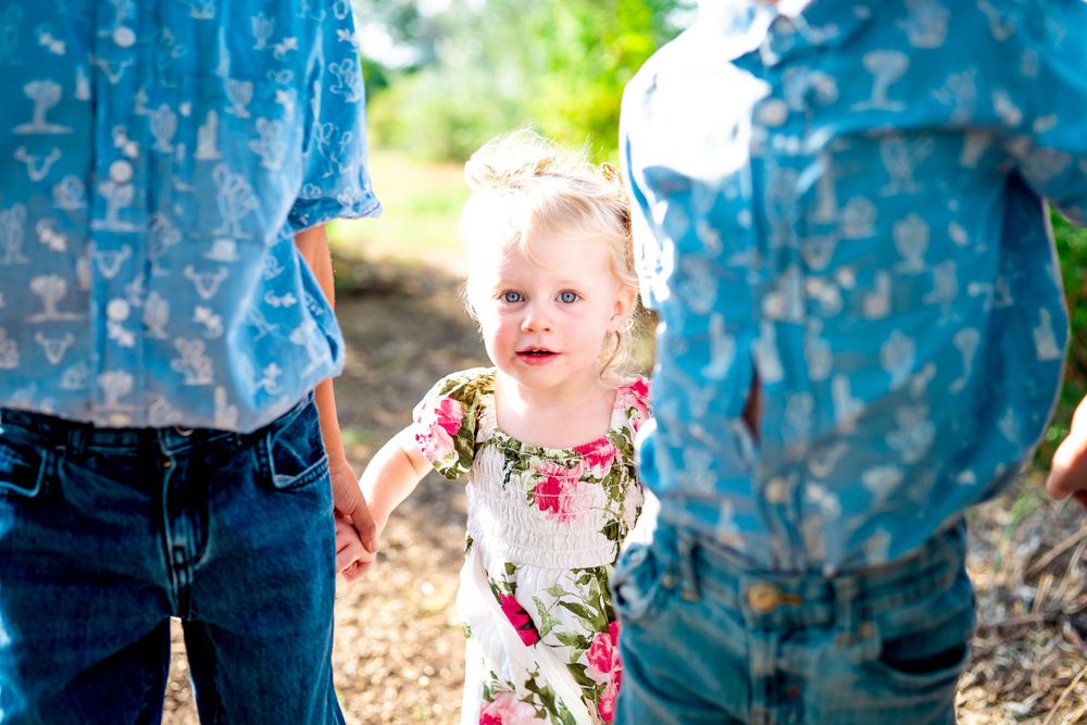 Child in floral dress walking between adults wearing blue shirts outdoors.